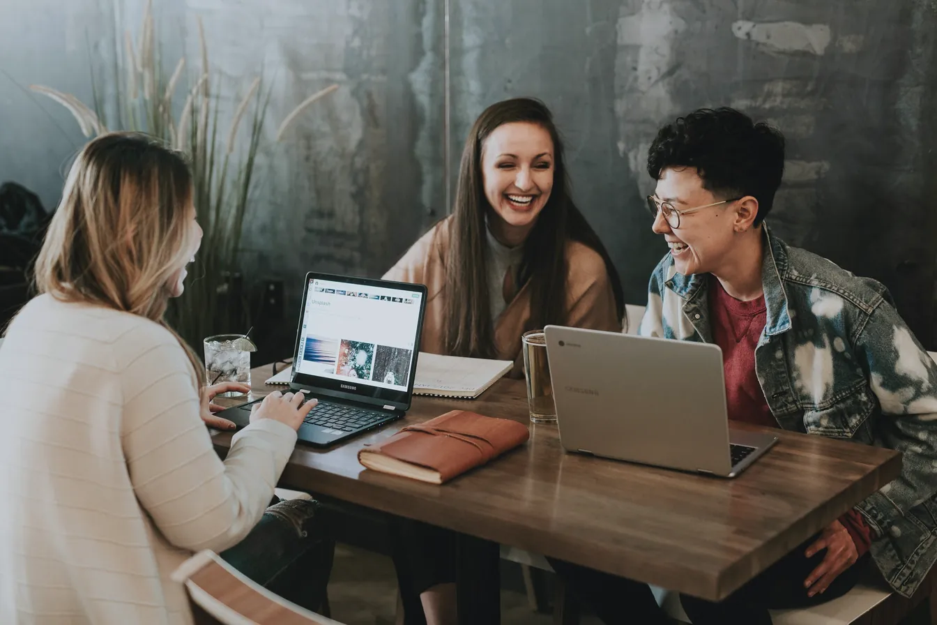 Trois personnes échangent en souriant autour d'une table, ordinateurs ouverts.